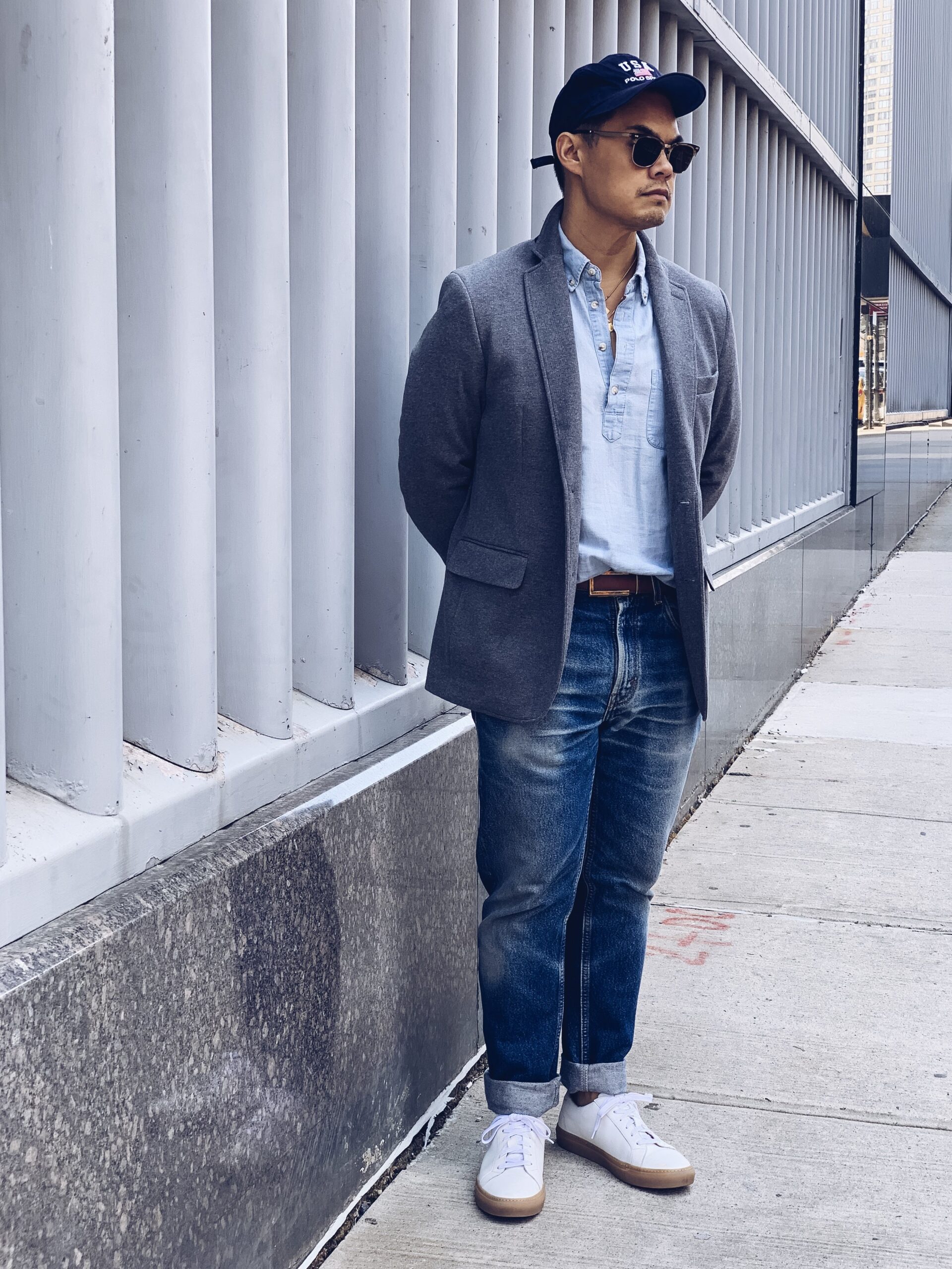 man wearing white sneakers with blue jeans light blue button down shirt and grey sportcoat with blue baseball cap