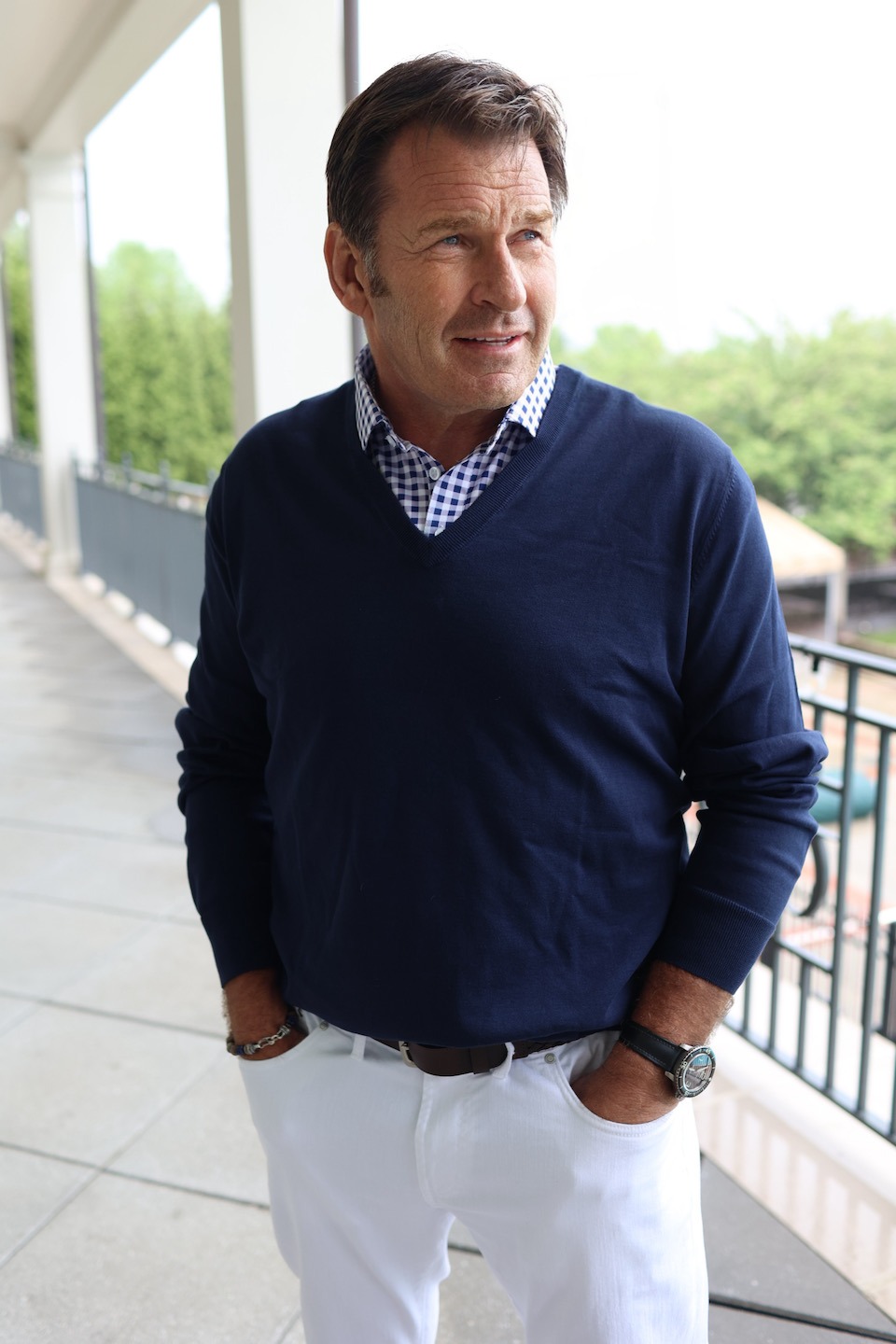 man wearing blue gingham shirt under navy v neck sweater and white jeans