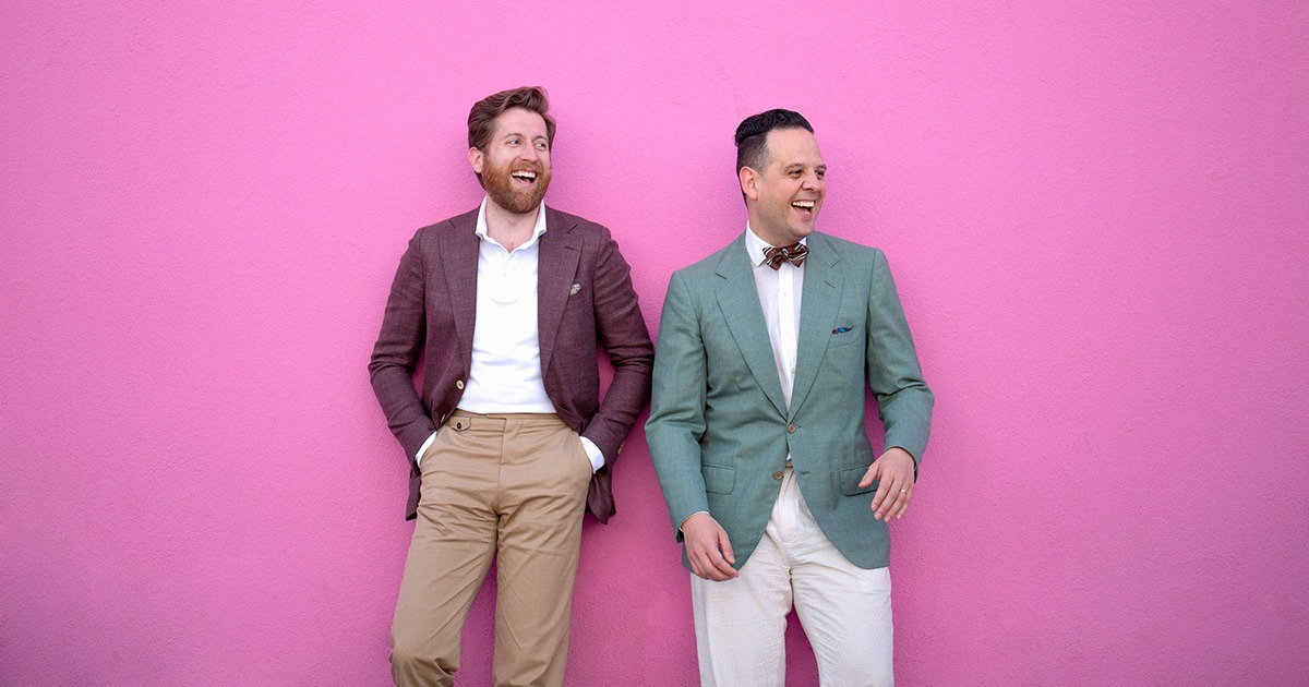 two man in suit jackets standing against a pink wall