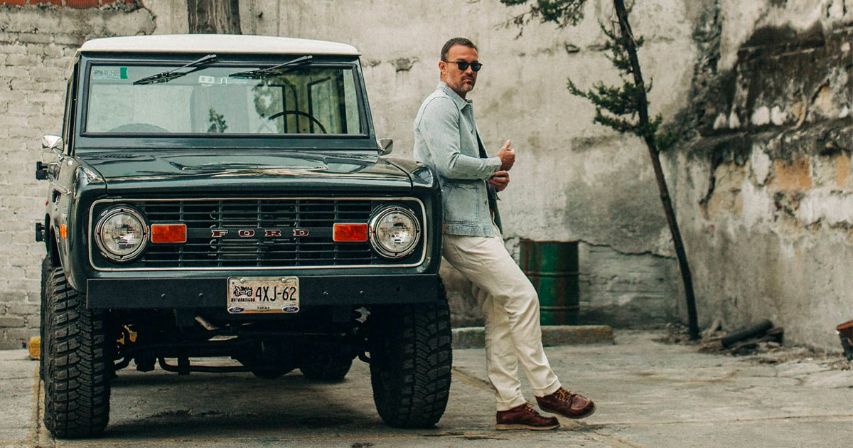 man wearing white jeans and a light blue jacket leaning against an old ford suv